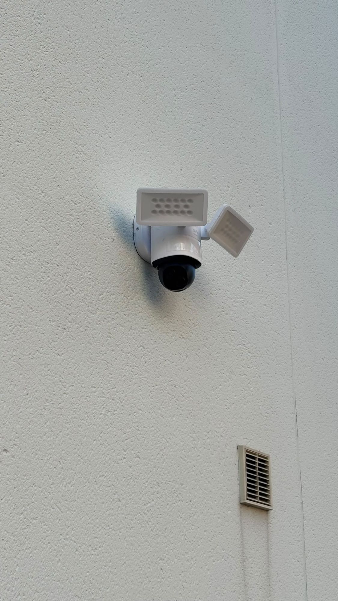 A white floodlight camera fitted high on a rendered wall