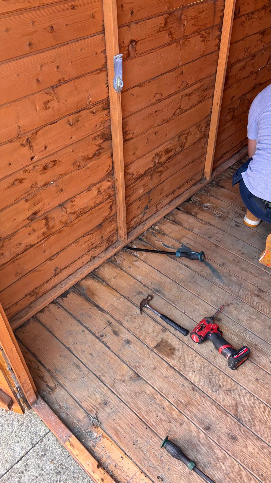 Inside the shed during the repair, with a pry bar, hammer and drill on the floor and Spiros kneeling at the side